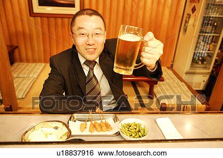 Stock Photograph - Portrait of mature man sitting at bar counter, raising glass of beer. Fotosearch