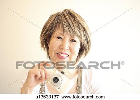 Stock Photo - Portrait of mature woman holding digital camera, smiling, white background. Fotosearch