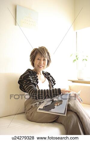 Stock Photo - Portrait of mature woman sitting on sofa, holding magazine. Fotosearch