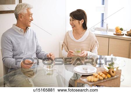 Stock Image - Senior couple drinking tea at dining table, smiling. Fotosearch