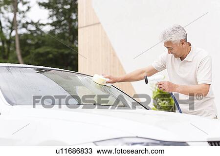 Stock Image - Senior man washing car, holding hose and sponge. Fotosearch