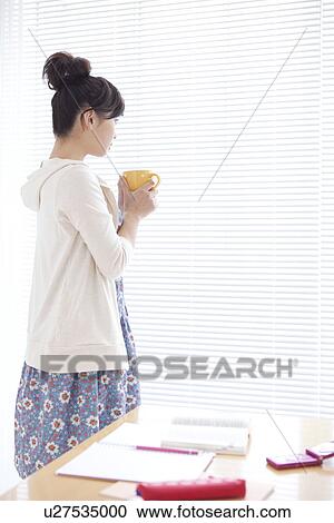 Stock Image - Young woman holding coffee and looking out the window. Fotosearch