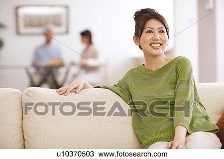 Young woman sitting on sofa, parents in background, differential focus View Large Photo Image Stock Image - Young woman sitting on sofa, parents in background, differential focus. Fotosearch