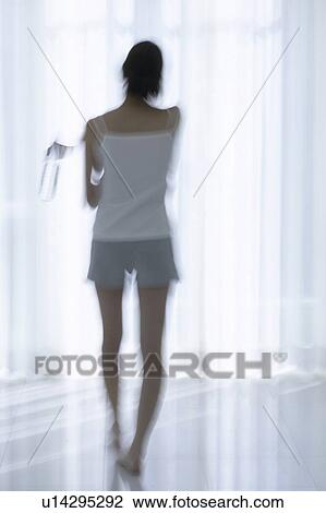 Stock Image - Young woman standing by window, holding bottle of water, soft focus, blurred motion. Fotosearch