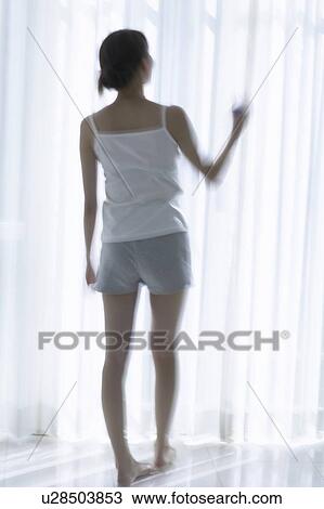 Stock Image - Young woman standing by window. Fotosearch