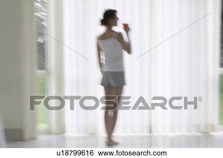Stock Photograph - Young woman standing by window, drinking glass of juice, soft focus, blurred motion. Fotosearch