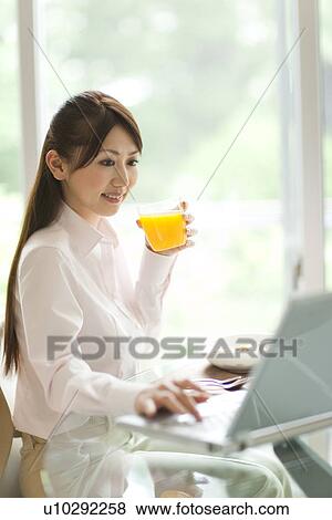Stock Photo - Young woman using laptop at table, holding glass of orange juice. Fotosearch