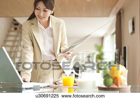Stock Photography - Young woman using laptop at table, holding newspaper. Fotosearch