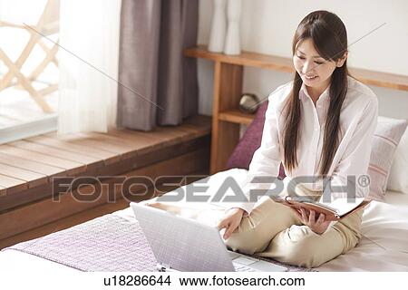 Picture - Young woman using laptop on bed, holding personal organizer. Fotosearch