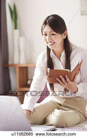 Stock Photography - Young woman using laptop on bed, holding personal organizer. Fotosearch