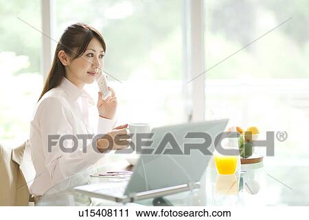Stock Image - Young woman using mobile phone at table, holding mug. Fotosearch