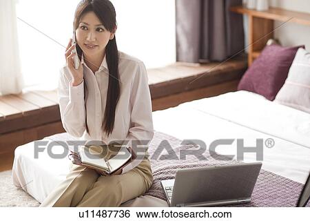 Stock Photograph - Young woman using mobile phone on bed, holding personal organizer. Fotosearch