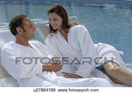 Stock Photo - Couple in bathrobes is lying next to the swimming pool. Fotosearch