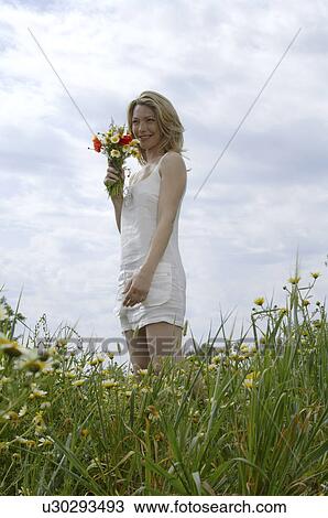 Stock Image - White dressed women is standing with a bunch of flowers in a meadow. Fotosearch