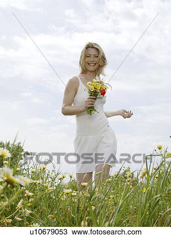 Stock Image - White dressed women with a bunch of flowers is running through a meadow. Fotosearch