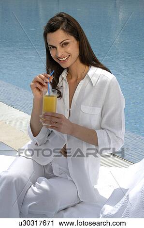 Stock Image - Woman dressed in white enjoying a drink at the swimming pool. Fotosearch
