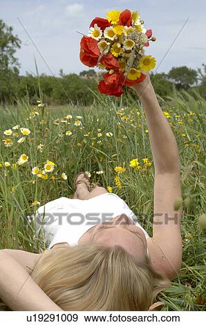 Stock Photo - Woman is lying in a meadow and holding a bunch of flowers in the air. Fotosearch