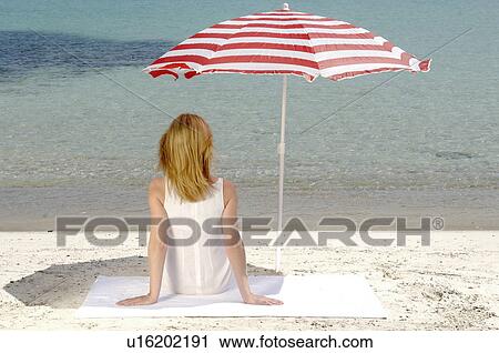 Stock Image - Woman sitting on the beach under a parasol - back view. Fotosearch