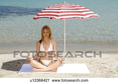 Stock Photo - Woman sitting on the beach under a parasol - front view. Fotosearch