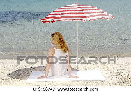 Stock Photo - Woman sitting on the beach under a parasol - back view. Fotosearch