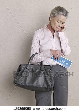 Stock Image - Senior citizen in business wear with ticket and carpetbag using a mobile. Fotosearch