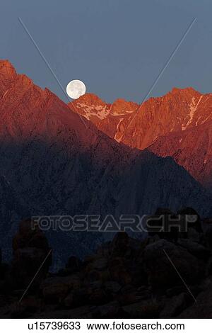 Stock Image - Full moon setting over snow-tipped High Sierras mountains, with red light of dawn lighting the range. Fotosearch