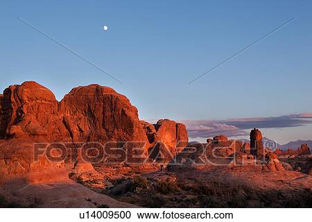 Stock Image - Garden of Eden, Arches at sunset, with rock glowing red in setting sun, full moon rising in blue sky, with pink clouds forming, Utah. Fotosearch