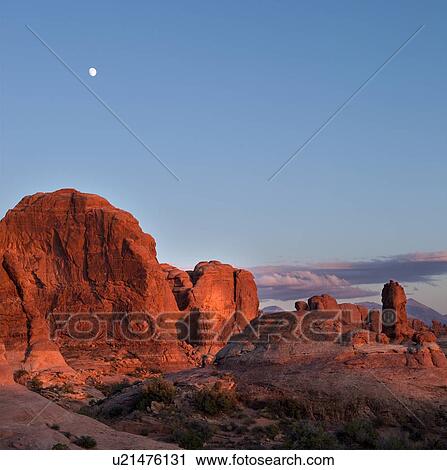 Stock Image - Garden of Eden, Arches at sunset, with rock glowing red in setting sun, full moon rising in blue sky, with pink clouds forming, Utah. Fotosearch