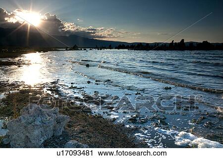 Stock Image - Gentle waves and foam of Mono Lake shoreline, sunlit in summer evening light, with sunstar behind looming clouds. Fotosearch