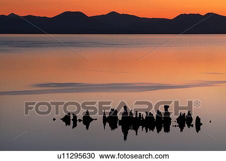 Stock Image - Orange dawn light silhouettes the tufa formations of Mono Lake, with mountain range beyond, on a calm summer morning, with deep reflections in the still water. Fotosearch