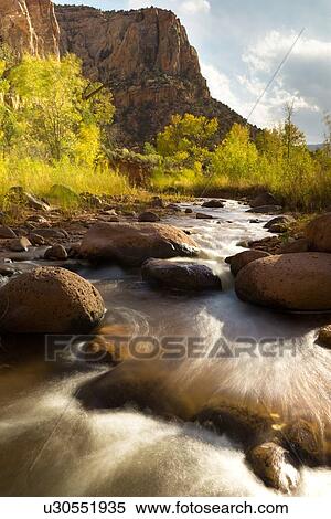 Stock Photography - Pleasant Valley, river with aspens alongside, steep rocky slope to mountains, blue sky, white puffy clouds, water motion slowed over rocks, Utah. Fotosearch