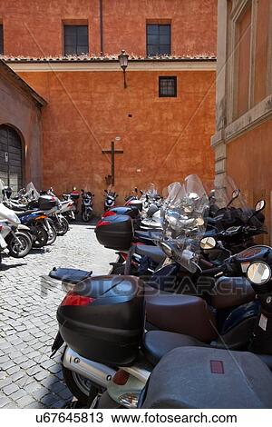 Stock Image - Row of motorbikes parked on sidestreet with orange stucco walls of church in background. Fotosearch