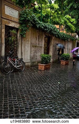 Stock Image - Via Magutta, cobblestone street, pedestrians in rain, Rome. Fotosearch