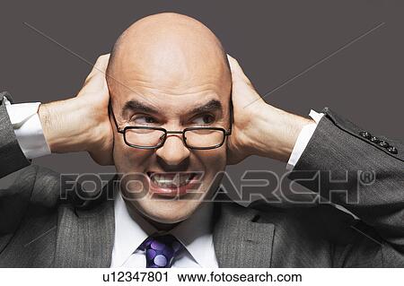 Stock Image - Bald businessman holding hand over ears, looking sideways, making a face. Fotosearch