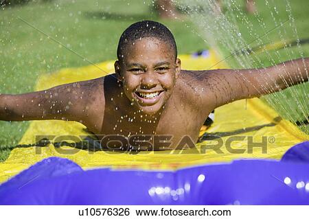 Stock Photograph - Boy (10-12) Sliding on water slide, front view portrait.. Fotosearch
