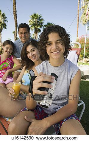 Stock Image - Boy (13-15) holding camcorder, sitting outdoors with sister (7-9) and parents, portrait.. Fotosearch