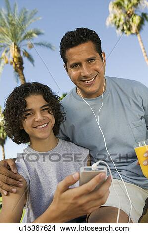 Picture - Boy (13-15) holding portable music player, father listening with earphones and holding glass of juice, front view portrait.. Fotosearch