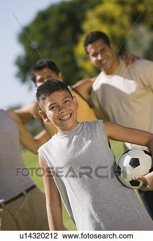 Stock Image - Boy (13-15) holding soccer ball, with three men in park.. Fotosearch