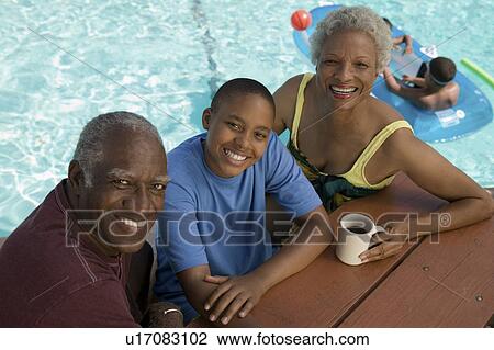 Stock Image - Boy (13-15) sitting with grandparents at picnic table by swimming pool, elevated view portrait.. Fotosearch