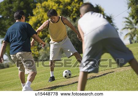 Stock Photo - Boy (13-15) with two young men, playing soccer outdoors in park.. Fotosearch