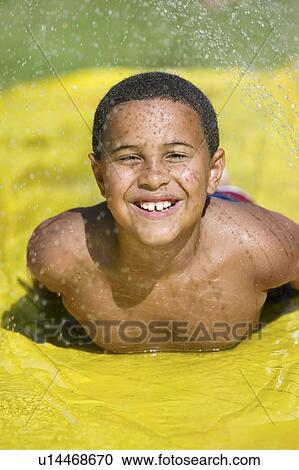 Stock Image - Boy (7-9) sliding on water slide, front view portrait.. Fotosearch