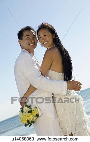 Stock Image - Bride and Groom embracing on beach, (portrait). Fotosearch