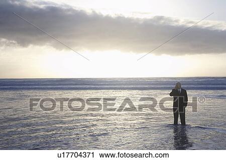 Stock Image - Business man using mobile phone standing in sea at sunset, elevated view. Fotosearch