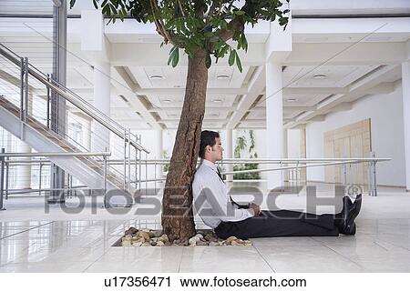 Stock Image - Businessman resting under tree outside office. Fotosearch