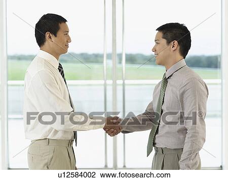 Stock Image - Businessmen standing, Shaking Hands in front of window, profile. Fotosearch