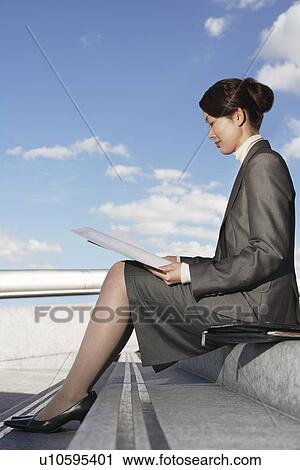 Stock Image - Businesswoman sitting outdoor steps, reading newspaper, profile. Fotosearch