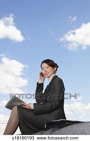 Stock Image - Businesswoman sitting outdoors, Using Cell Phone, reading newspaper, low angle view. Fotosearch
