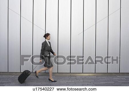 Businesswoman walking outdoors, pulling suitcase behind her, side view View Large Photo Image Stock Photo - Businesswoman walking outdoors, pulling suitcase behind her, side view. Fotosearch