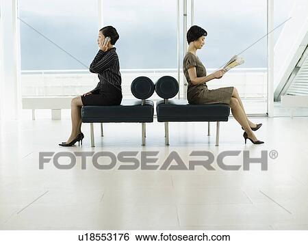 Stock Photograph - Businesswomen Sitting on Benches, facing opposite directions, talking on mobile, reading newspaper, profile. Fotosearch