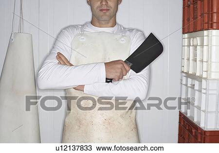Stock Image - Butcher standing, arms crossed, Holding Cleaver next to stack of crates, mid section. Fotosearch